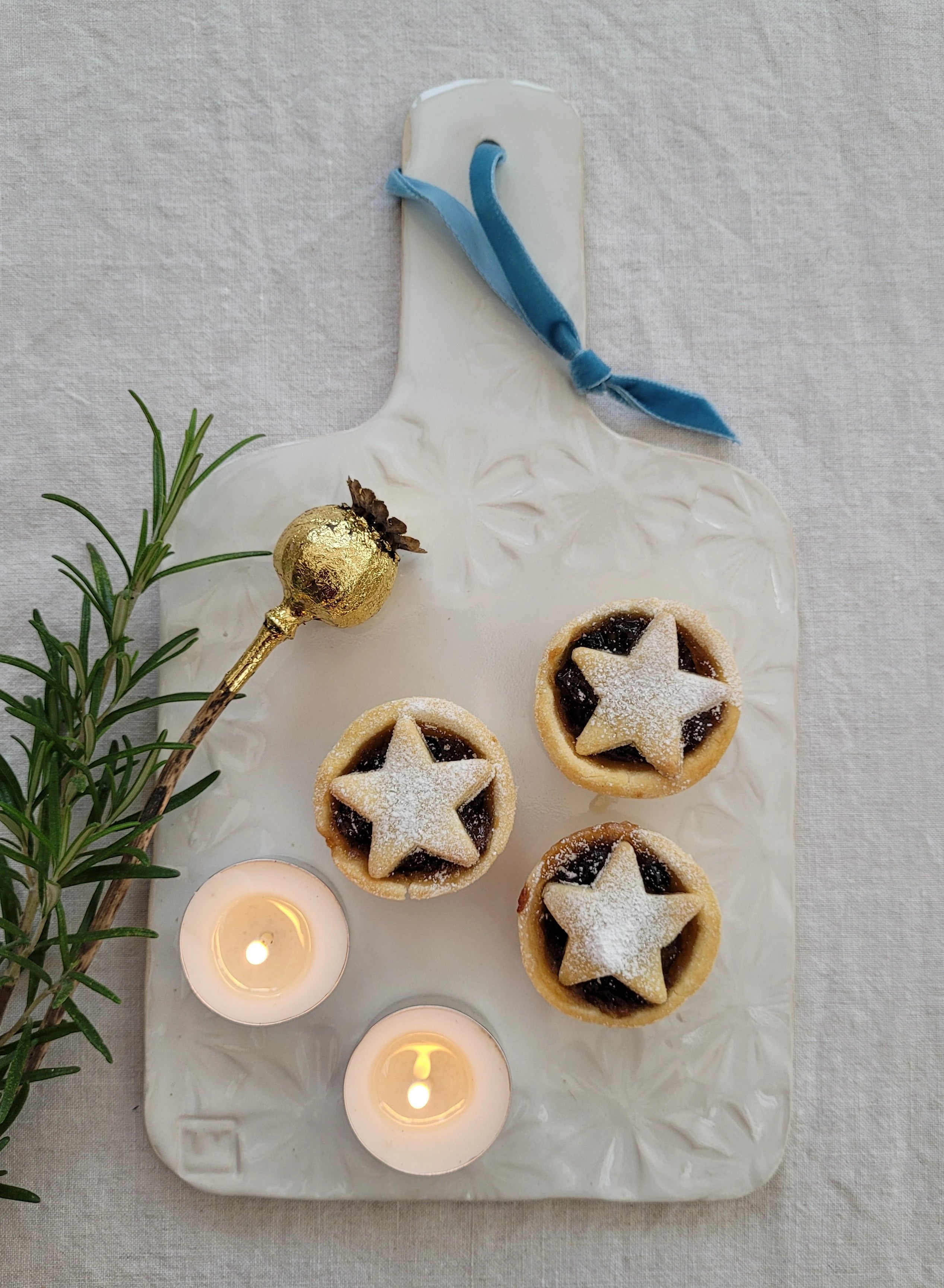 Handmade ceramic cutting board made by lizzie mabley, set with mince pies, candles, and a gold bell on a light background