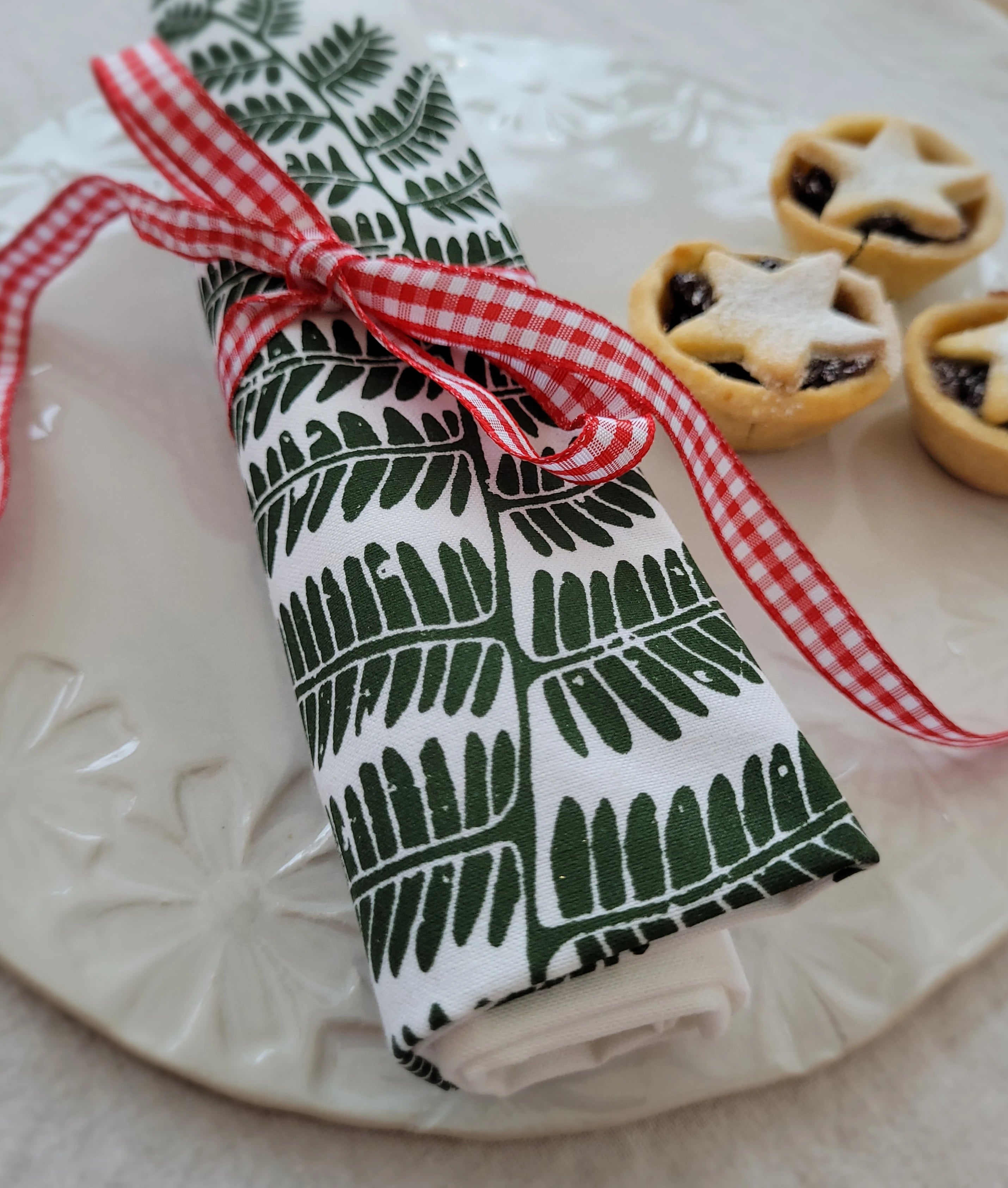 Lizzie Mabley designed fern napkin wrapped in red and white checkered ribbon on a handmade ceramic plate with mince pies.